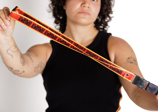 Clothing The Gaps. Power Lanyard. Thick strap with black 'always was' text on a red background with a yellow trim repeated. With an extra large silver metal clasp and engraved with the Clothing The Gaps logo. Black safety clip at back of neck. Silver metal clip at end of lanyard to attach what you would like.