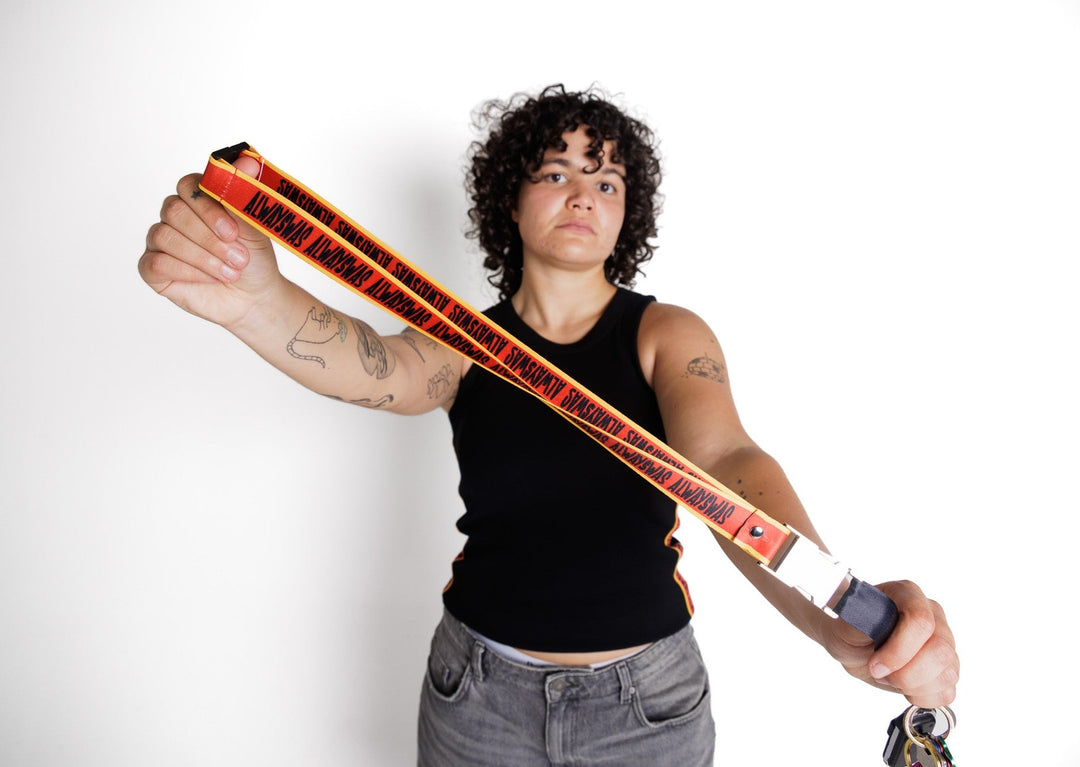 Clothing The Gaps. Power Lanyard. Thick strap with black 'always was' text on a red background with a yellow trim repeated. With an extra large silver metal clasp and engraved with the Clothing The Gaps logo. Black safety clip at back of neck. Silver metal clip at end of lanyard to attach what you would like.