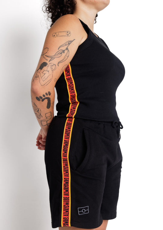 Clothing the Gaps. Black cotton shorts. With black drawstrings. Embroiled black outlined aboriginal flag in corner. Black 'always was always will be' text on a red background with a yellow trim. In a cotton tape strip going down both sides of shorts.