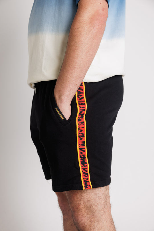Clothing the Gaps. Black cotton shorts. With black drawstrings. Embroiled black outlined aboriginal flag in corner. Black 'always was always will be' text on a red background with a yellow trim. In a cotton tape strip going down both sides of shorts.