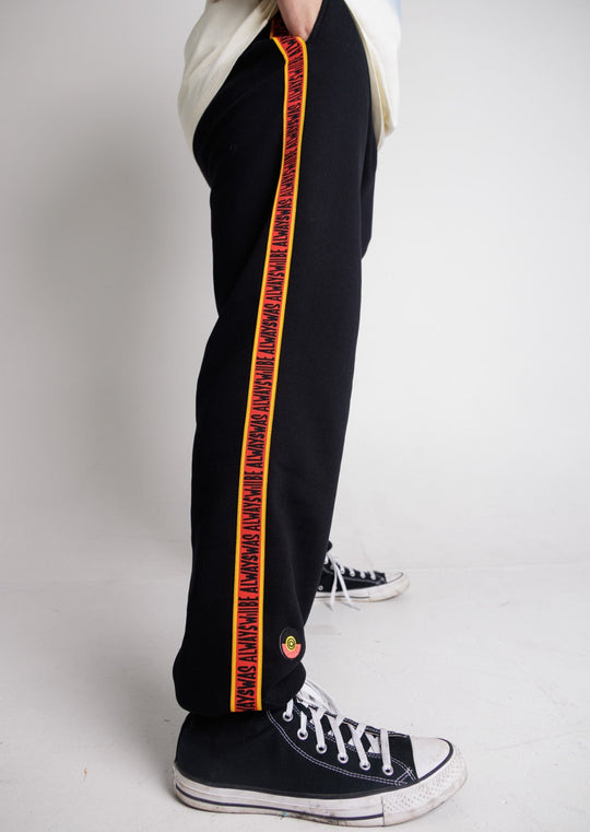 Clothing the Gaps. Black cotton Pants. With black drawstrings. Elastic cuts and waistband. Embroiled Clothing The Gaps logo. Black 'always was always will be' text on a red background with a yellow trim. In a cotton tape strip going down both sides of pants.