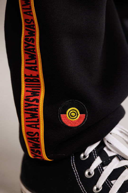 Clothing the Gaps. Black cotton Pants. With black drawstrings. Elastic cuts and waistband. Embroiled Clothing The Gaps logo. Black 'always was always will be' text on a red background with a yellow trim. In a cotton tape strip going down both sides of pants.