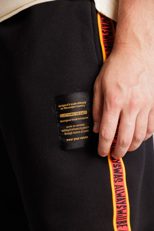 Clothing the Gaps. Black cotton Pants. With black drawstrings. Elastic cuts and waistband. Embroiled Clothing The Gaps logo. Black 'always was always will be' text on a red background with a yellow trim. In a cotton tape strip going down both sides of pants.