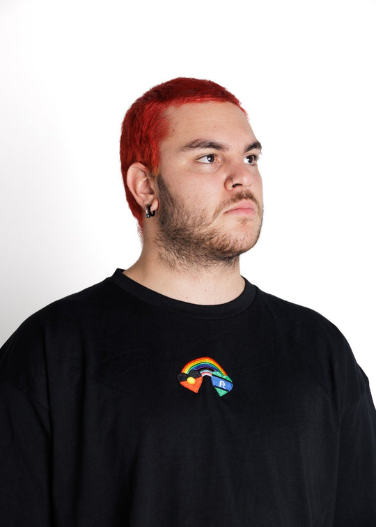 Clothing The Gaps. Blak Rainbow Luv Crop. All black boxy crop tee with  a heart featuring the Aboriginal flag at the start of a rainbow and at the end of the rainbow the Torres Strait Islander flag. This design is embroidered on the front of the t-shirt. The rainbow includes the colours from the progress pride flag. Show your pride or support for First Nations LGBTQIAS&B+ Community.