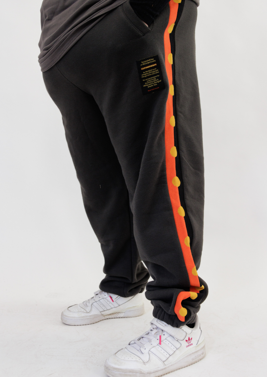 Clothing The Gaps. Charcoal Flag Track Pants. Charcoal dark grey track pants with Aboriginal flag tape down the sides. Has a elastic waist with charcoal grey draw strings and cuffed ankles.