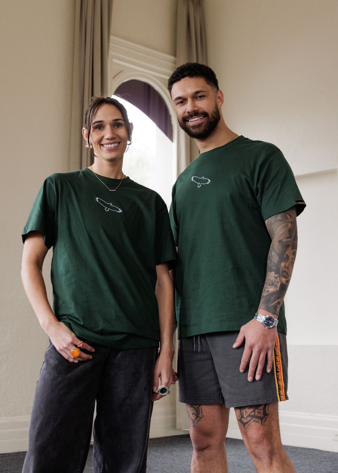 Two people wearing green bunjil Clothing The Gaps t-shirts standing indoors with a neutral background