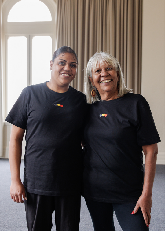 Two women wearing black Blak Luv Clothing The Gaps t-shirts with colorful designs standing indoors.