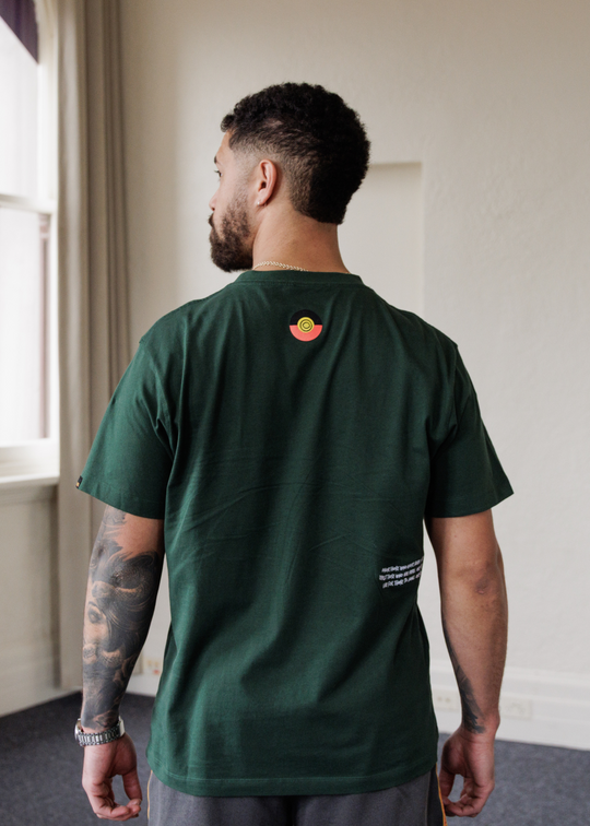 Man wearing a green  Bunjil eagle clothing the gaps t-shirt with a logo on the back, standing in a room with a window.