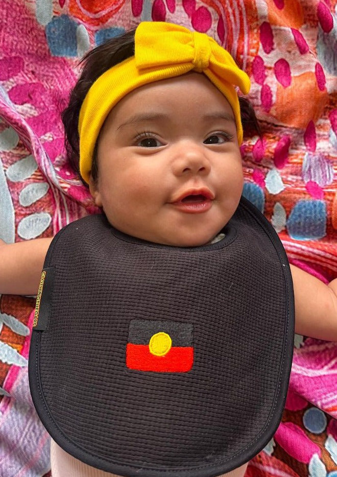 Clothing The Gaps. Flag Bubup Baby Bibs. Black bib cotton waffle material with red, black and yellow Aboriginal flag embroidered in centre. Includes Velcro to attach around babies neck.
