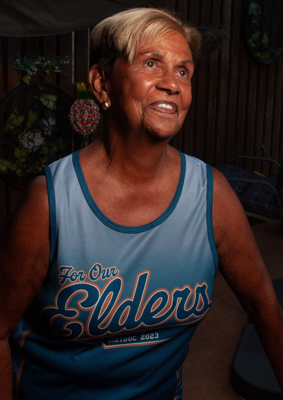 Clothing The Gaps. For Our Elders Training Singlet.  Polyester blue gradient singlet with dark blue trim around arms and neck. 'For Our Elders NAIDOC 2023' text in dark blue in centre of shirt outlined with a light brown and white. Down the sides in a light contrasting blue under the arms artwork done by Gunditjmara woman, Laura Thompson.