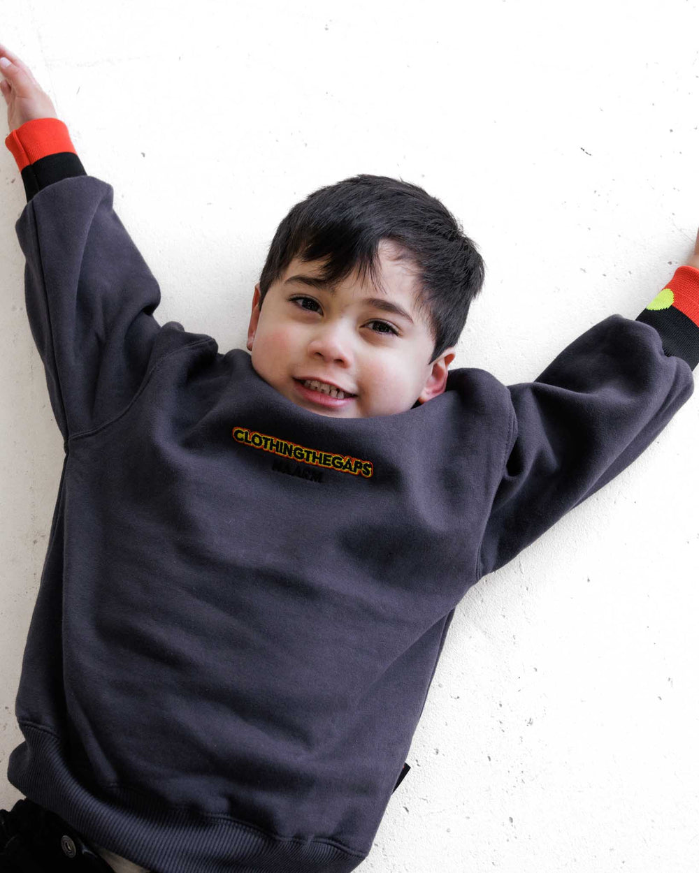 Clothing The Gaps. Kids Charcoal Crew Jumper. Charcoal dark grey crew neck jumper. With black, yellow and red 'Clothing the gaps' and the word 'Narrm' embroidered on the front of crewneck.' The wrist cuffs of the crew have a woven aboriginal flag on them. 