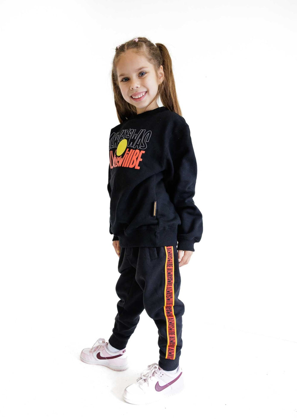 Clothing the Gaps. KIDS 'Always Was, Always Will Be' Track Pants. Black cotton Pants. With black drawstrings. Elastic cuts and waistband. Embroiled Clothing The Gaps logo. Black 'always was always will be' text on a red background with a yellow trim. In a cotton tape strip going down both sides of pants. 