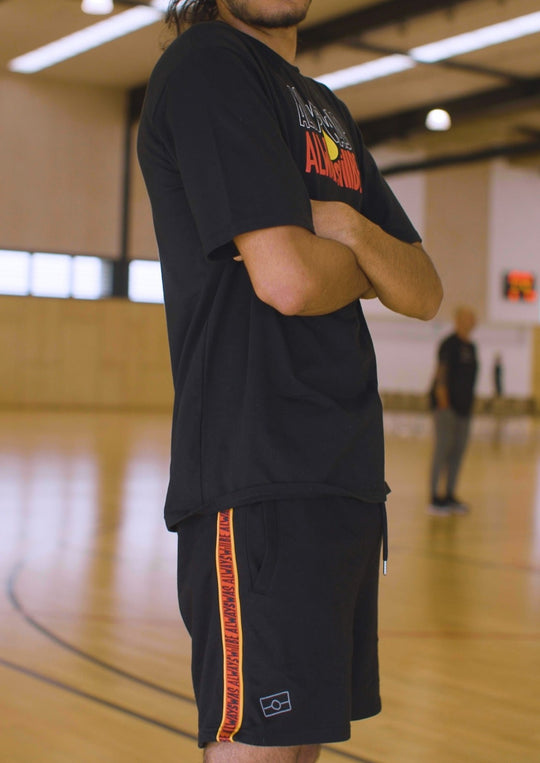 Clothing the Gaps. Black cotton shorts. With black drawstrings. Embroiled black outlined aboriginal flag in corner. Black 'always was always will be' text on a red background with a yellow trim. In a cotton tape strip going down both sides of shorts.