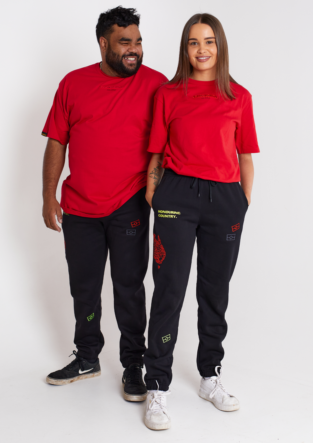 Clothing The Gaps. Honouring country track pants. Black warm track pants. With embroidered decolonised map of 'Australia' in red on side of right knee and above the embroidered phrase 'Honour Country' in bright yellow on middle of right thigh. Embroidered flags bright yellow at bottom of right leg and a red and grey flag on pocket of left leg. Features deep side pockets, elastic waist with black draw strings.