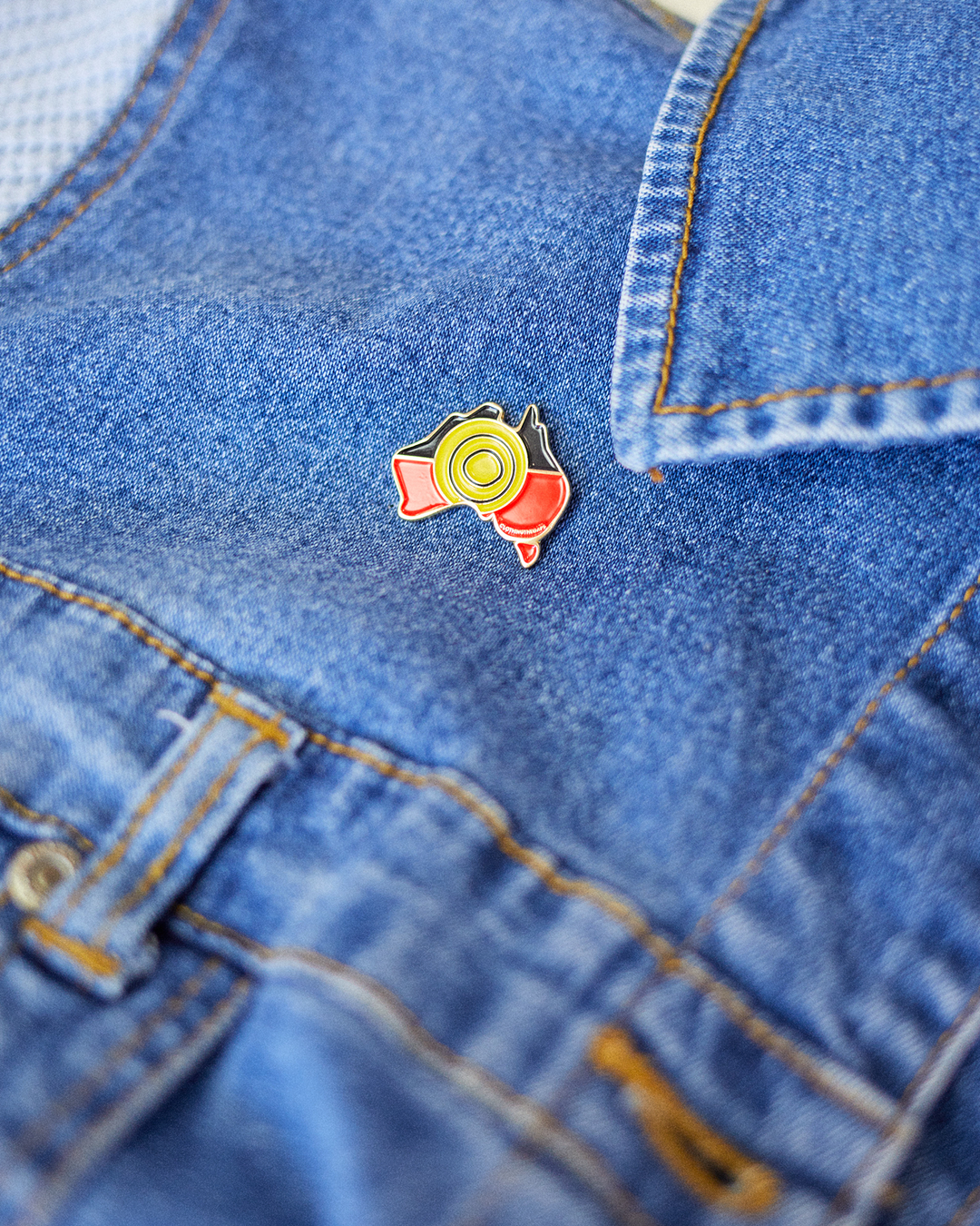 Clothing The Gaps. Pin. Black, yellow and red Aboriginal flag colours shaped inside map of Australia with gold outline and gold backing.