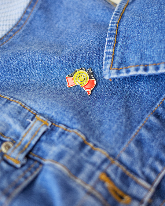 Clothing The Gaps. Pin. Black, yellow and red Aboriginal flag colours shaped inside map of Australia with gold outline and gold backing.