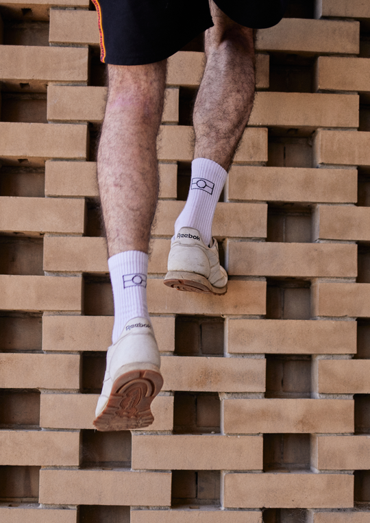 Clothing The Gaps. White Empty Flag Socks. Long white socks with black Aboriginal flag outline on back. Black on bottom of sock to hide dirt. Clothing The Gaps written above toe area.