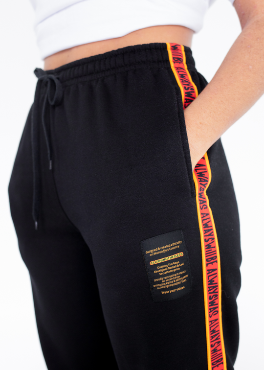 Clothing the Gaps. Black cotton Pants. With black drawstrings. Elastic cuts and waistband. Embroiled Clothing The Gaps logo. Black 'always was always will be' text on a red background with a yellow trim. In a cotton tape strip going down both sides of pants.