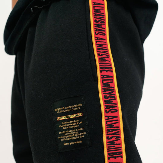 Clothing the Gaps. Black cotton Pants. With black drawstrings. Elastic cuts and waistband. Embroiled Clothing The Gaps logo. Black 'always was always will be' text on a red background with a yellow trim. In a cotton tape strip going down both sides of pants.