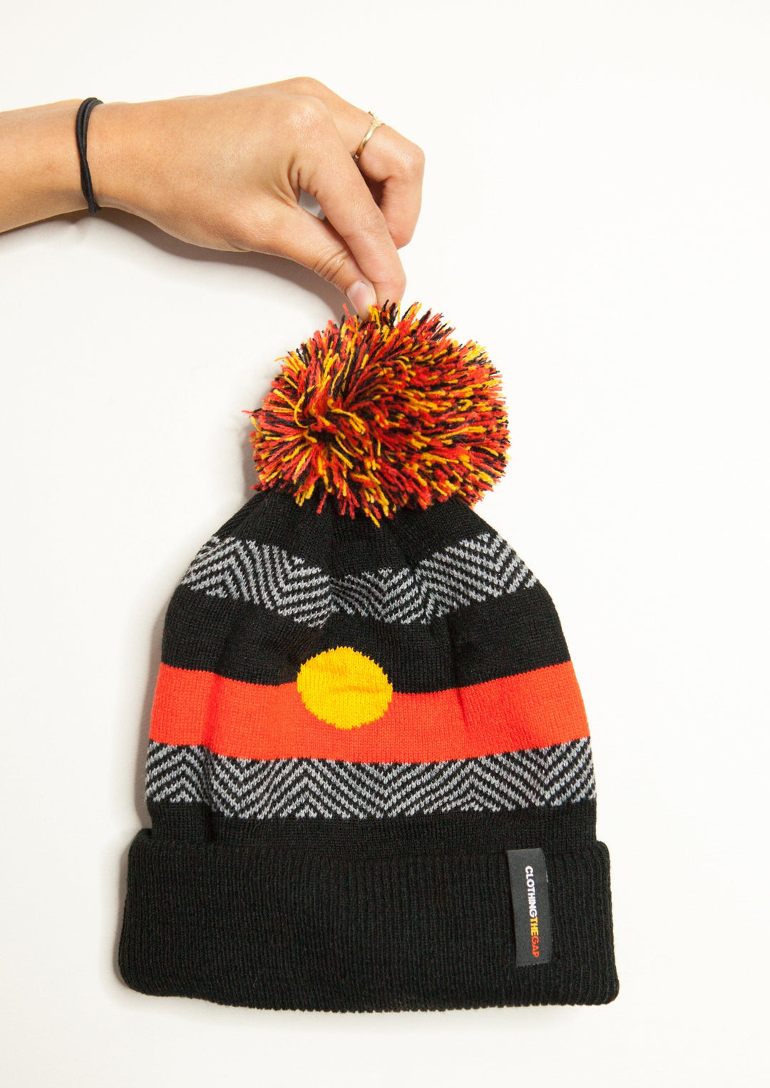 Clothing the Gaps. Beanie with Aboriginal flags on both sides and contrasting black and dark grey line work and red, black and yellow pom pom on top.