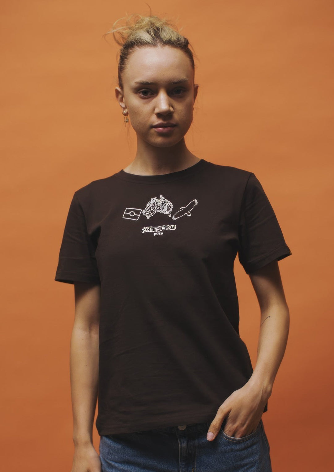 Clothing The Gaps. Icon Tee. Expresso dark brown tee featuring Aboriginal symbols of resistance and culture including a white outline of the Aboriginal flag, a white outlined decolonised map of “Australia” and a white outlined Bunjil, a well known wedge-tailed eagle - who is the spiritual creator and protector of the Kulin Nation in Victoria. These symbols are on the front of the tee across the chest.