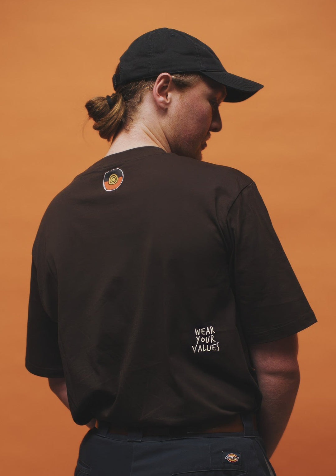 Clothing The Gaps. Icon Tee. Expresso dark brown tee featuring Aboriginal symbols of resistance and culture including a white outline of the Aboriginal flag, a white outlined decolonised map of “Australia” and a white outlined Bunjil, a well known wedge-tailed eagle - who is the spiritual creator and protector of the Kulin Nation in Victoria. These symbols are on the front of the tee across the chest.