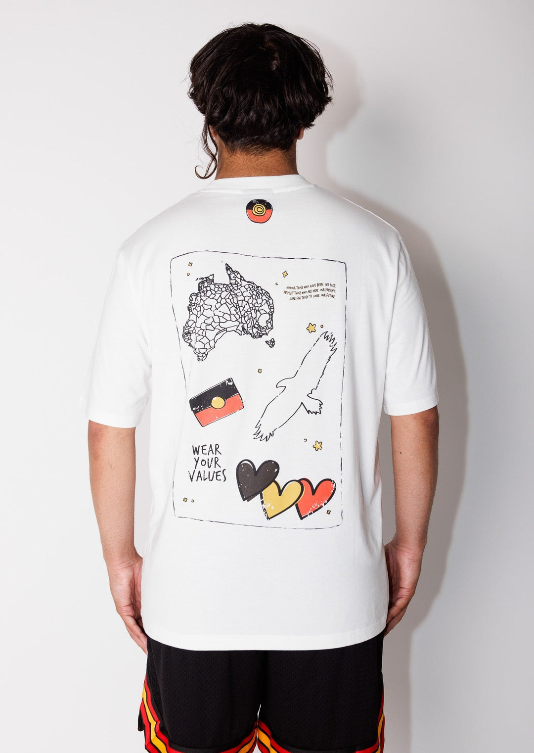 Clothing The Gaps. Icon Spirit Tee. White T-shirt with 'Clothing The Gaps' in a black, yellow, red handwritten text small on front of t-shirt in left pocket area. Back of tee features prominent Aboriginal symbols of resistance and culture, including the Aboriginal flag, a black outlined decolonised map of “Australia,” outlined black Bunjil, 3 black, yellow and red hearts and black handwritten text 'wear your values'