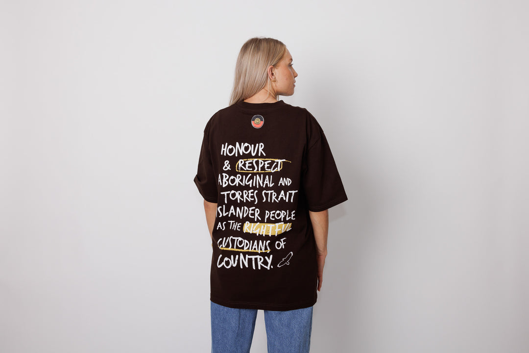 Clothing The Gaps. Handwritten Honour Country Tee. Expresso dark brown T-shirt with 'Honour Country' across chest in handwritten white text and coloured Aboriginal flag small underneath. On Back of tee white hand-writen style text 'Honour and respect Aboriginal and Torres Strait Islander people as the rightful custodians of country.' Circling 'respect' underlining 'custodians' and highlighting 'rightful' in a sand yellow colour. Outlined 'bunjil' eagle in bottom right corner.'    