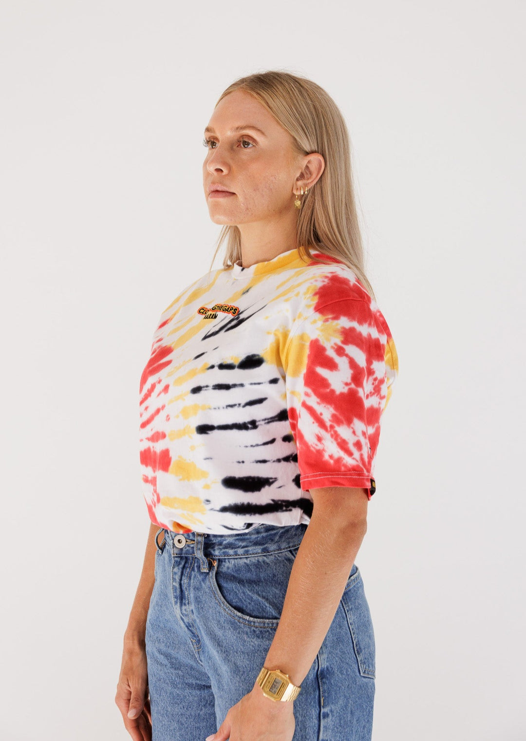 Clothing The Gaps. Tie Dye Spirit Tee. White T-shirt. With Red, black and yellow tie died in random pattern. Has embroidered 'Clothing The Gaps' on front chest with minimalist font in black text with a yellow and red outline and 'Narrm' in black embroidered underneath, acknowledging the land on which Clothing The Gaps operates it's social enterprise.