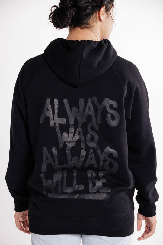 Clothing The Gaps. Black hoodie with Aboriginal flag outline embroidered in black on the front. On the back of the hoodie a powerful Aboriginal chant 'Always Was, Always Will Be' looks like it has been sprayed on with a black spray can! The Clothing the Gaps circle logo patch provides a sneaky little pop of red, black and yellow on the sleeve.