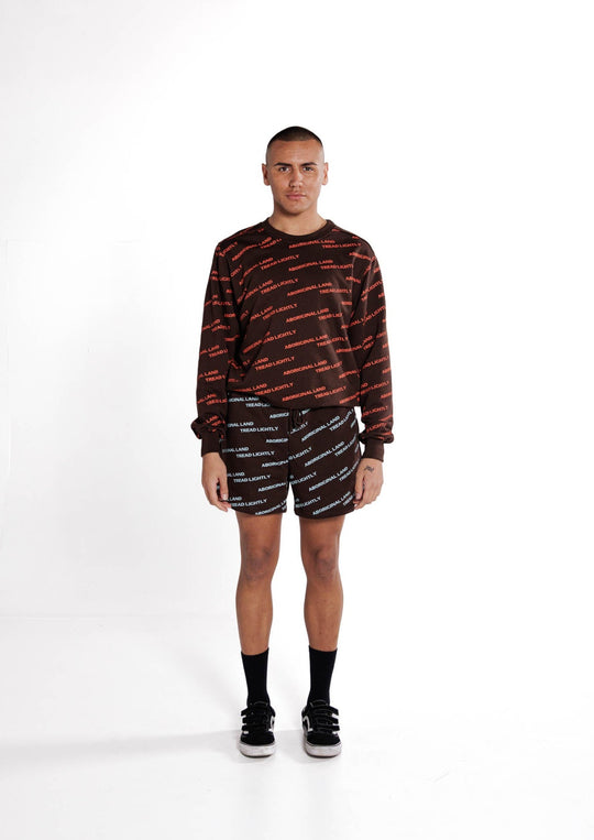 Clothing The Gaps. Orange Tread Lightly Crew. Brown Crewneck jumper with repeating pattern all over crew of the words 'Aboriginal Land Tread Lightly' text in a orange colour. Solid brown cuffs, waist band and neckline.