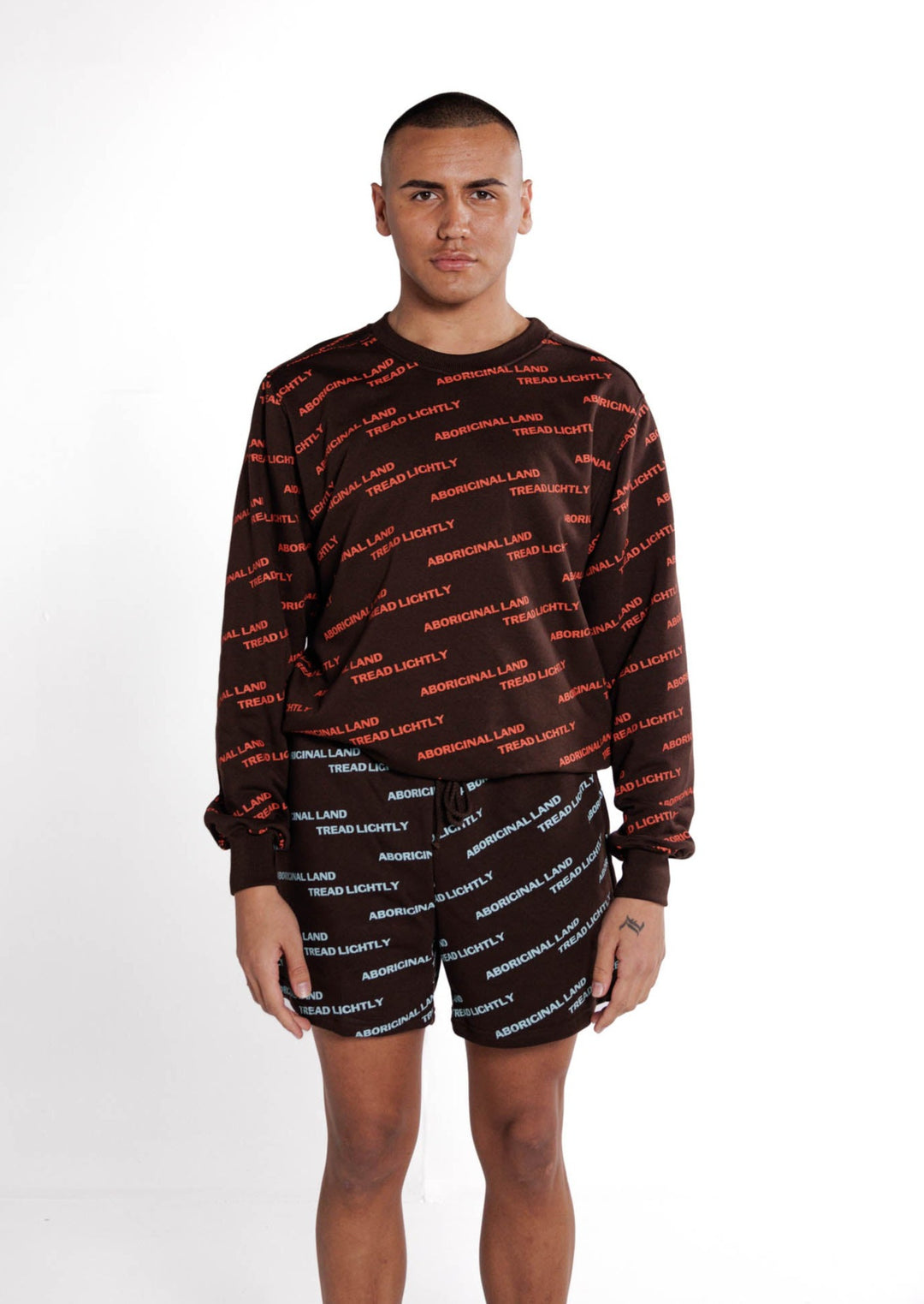 Clothing The Gaps. Orange Tread Lightly Crew. Brown Crewneck jumper with repeating pattern all over crew of the words 'Aboriginal Land Tread Lightly' text in a orange colour. Solid brown cuffs, waist band and neckline.