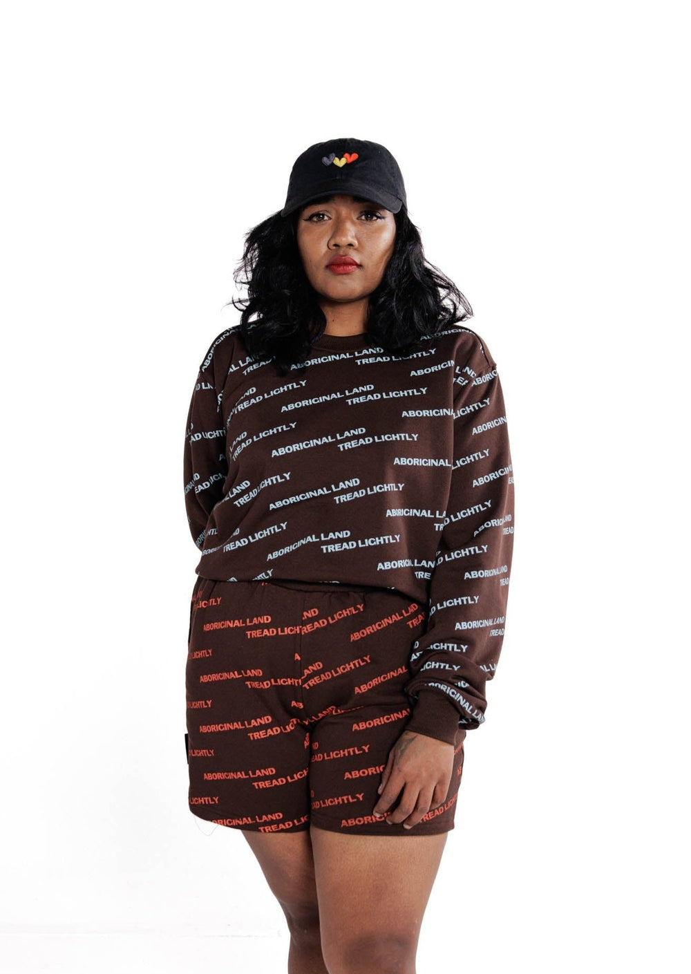 Clothing The Gaps. Blue Tread Lightly Crew. Brown Crewneck jumper with repeating pattern all over crew of the words 'Aboriginal Land Tread Lightly' text in a light blue colour. Solid brown cuffs, waist band and neckline.