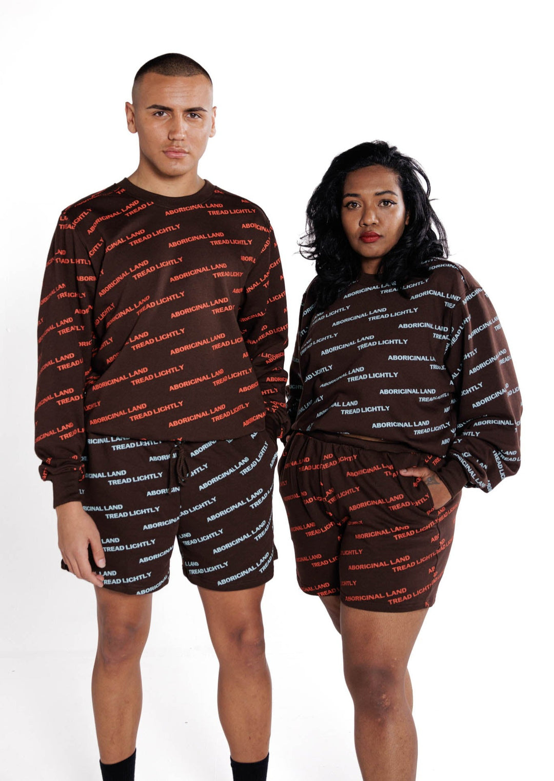 Clothing The Gaps. Orange Tread Lightly Crew. Brown Crewneck jumper with repeating pattern all over crew of the words 'Aboriginal Land Tread Lightly' text in a orange colour. Solid brown cuffs, waist band and neckline.