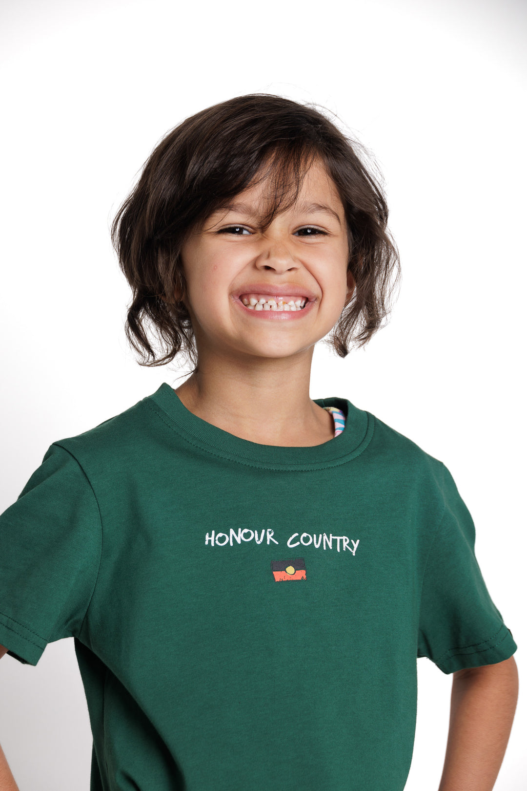 Clothing The Gaps. Kids Handwritten Honour Country Tee. Forrest green T-shirt with 'Honour Country' across chest in handwritten white text and coloured Aboriginal flag small underneath. On Back of tee white hand-writen style text 'Honour and respect Aboriginal and Torres Strait Islander people as the rightful custodians of country.' Circling 'respect' underlining 'custodians' and highlighting 'rightful' in a black colour. Outlined 'bunjil' eagle in bottom right corner.'        