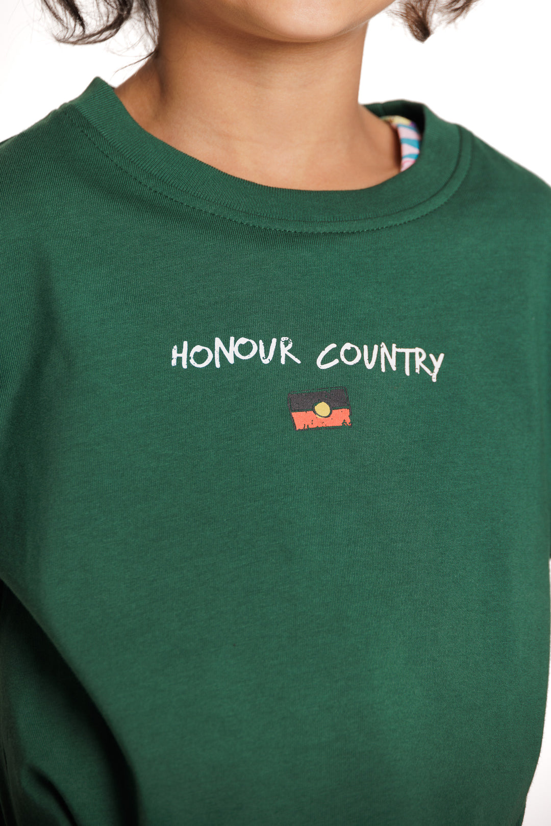 Clothing The Gaps. Kids Handwritten Honour Country Tee. Forrest green T-shirt with 'Honour Country' across chest in handwritten white text and coloured Aboriginal flag small underneath. On Back of tee white hand-writen style text 'Honour and respect Aboriginal and Torres Strait Islander people as the rightful custodians of country.' Circling 'respect' underlining 'custodians' and highlighting 'rightful' in a black colour. Outlined 'bunjil' eagle in bottom right corner.'        