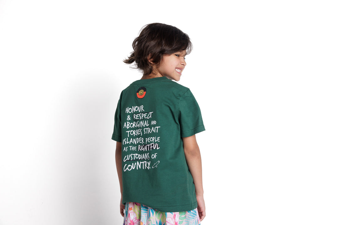 Clothing The Gaps. Kids Handwritten Honour Country Tee. Forrest green T-shirt with 'Honour Country' across chest in handwritten white text and coloured Aboriginal flag small underneath. On Back of tee white hand-writen style text 'Honour and respect Aboriginal and Torres Strait Islander people as the rightful custodians of country.' Circling 'respect' underlining 'custodians' and highlighting 'rightful' in a black colour. Outlined 'bunjil' eagle in bottom right corner.'        