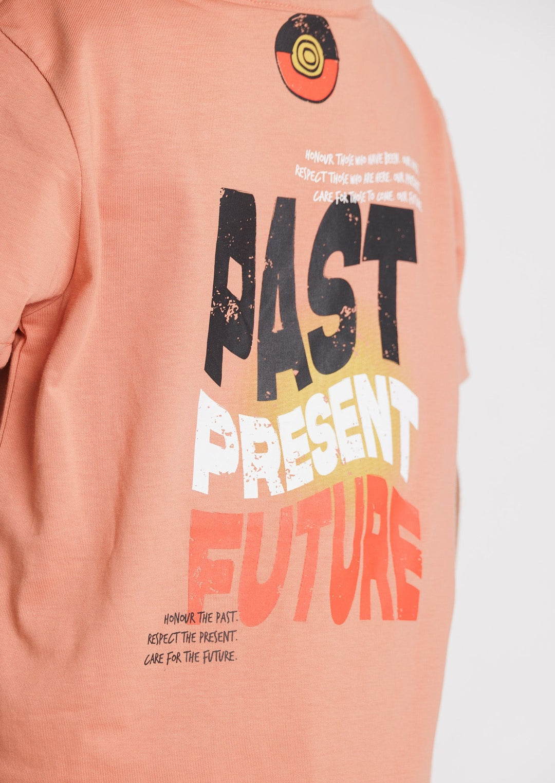 Clothing The Gaps. Kids Past Present Future Tee. Light peachy coloured tee with a small 'Past, Present, Future' pocket square on the front and large 'Past, Present , Future screen print back Black white and red with a yellow sun behind.