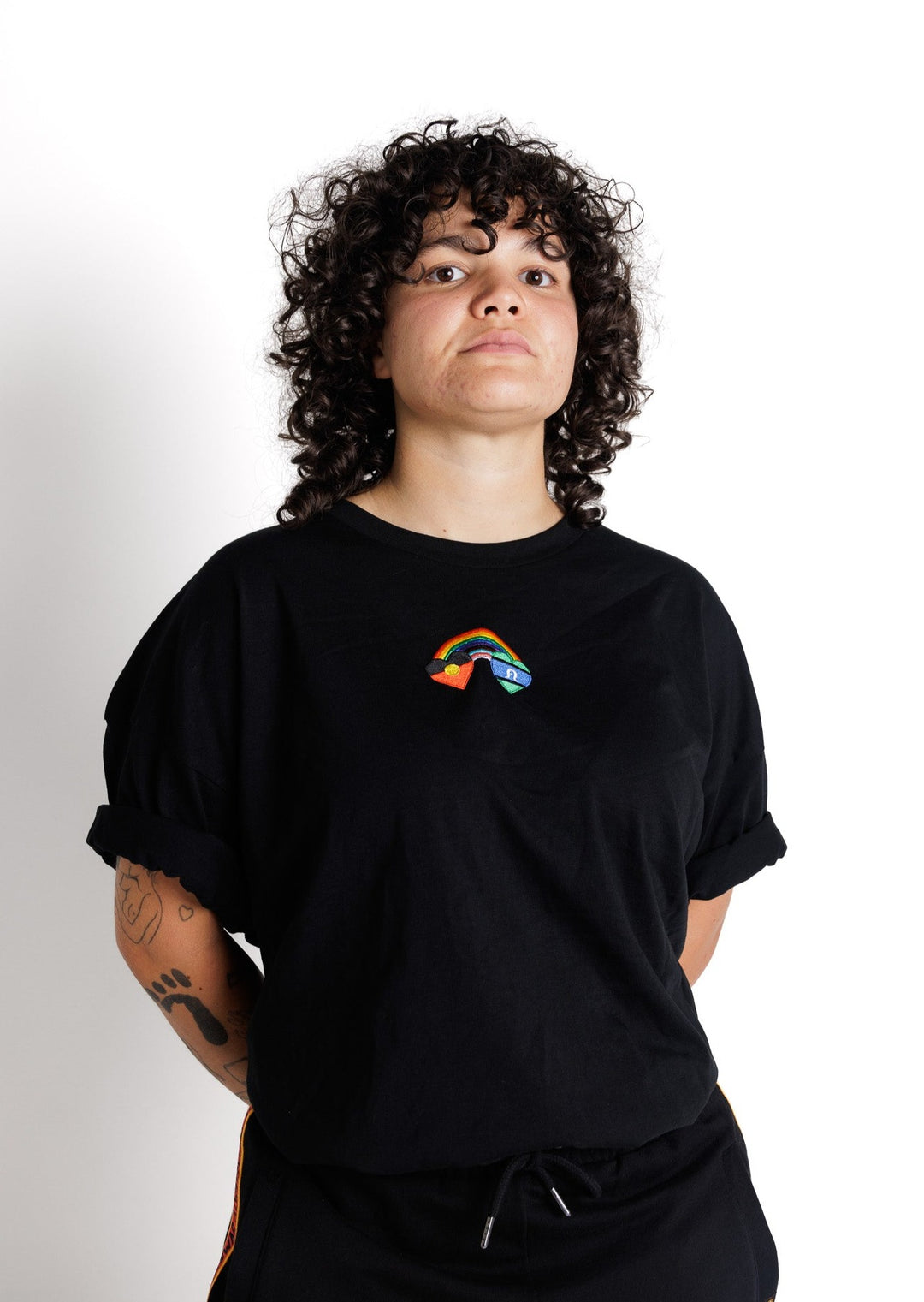 Clothing The Gaps. Blak Rainbow Luv Crop. All black boxy crop tee with  a heart featuring the Aboriginal flag at the start of a rainbow and at the end of the rainbow the Torres Strait Islander flag. This design is embroidered on the front of the t-shirt. The rainbow includes the colours from the progress pride flag. Show your pride or support for First Nations LGBTQIAS&B+ Community.