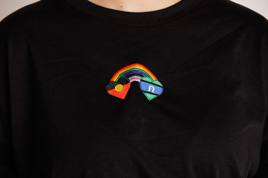 Clothing The Gaps. Blak Rainbow Luv Crop. All black boxy crop tee with  a heart featuring the Aboriginal flag at the start of a rainbow and at the end of the rainbow the Torres Strait Islander flag. This design is embroidered on the front of the t-shirt. The rainbow includes the colours from the progress pride flag. Show your pride or support for First Nations LGBTQIAS&B+ Community.