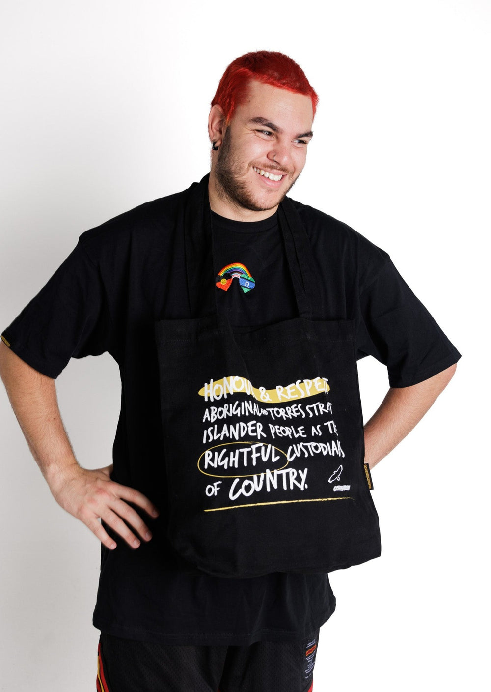 Clothing The Gaps. Honour Country Tote Bag. Black tote bag with 2 over the shoulder black straps. With white hand-writen style text 'Honour and respect Aboriginal and Torres Strait Islander people as the rightful custodians of country.' Circling 'rightful' underlining 'country' and highlighting 'honour and respect' in a sand yellow colour. Outlined 'bunjil' eagle in right corner.