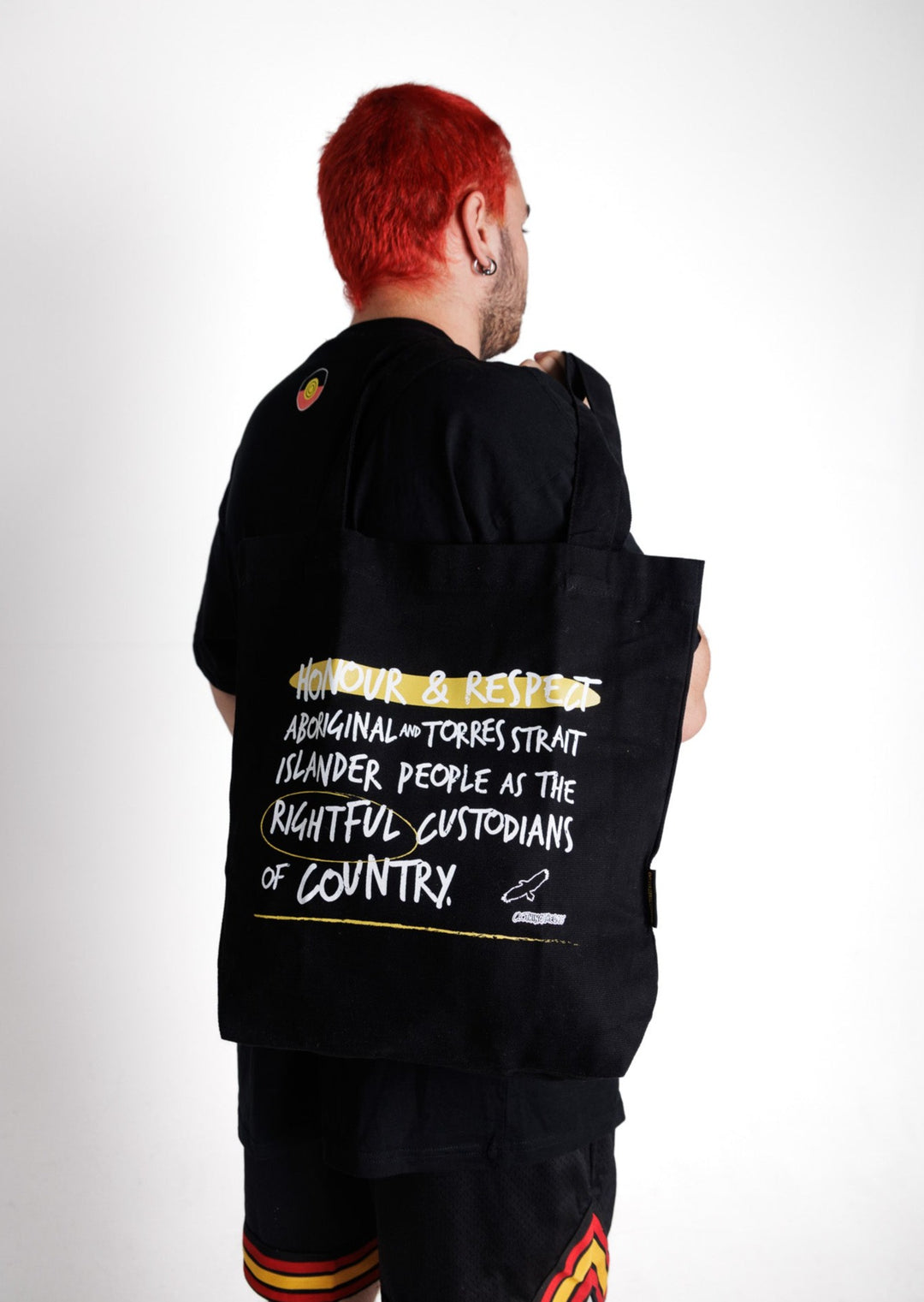 Clothing The Gaps. Honour Country Tote Bag. Black tote bag with 2 over the shoulder black straps. With white hand-writen style text 'Honour and respect Aboriginal and Torres Strait Islander people as the rightful custodians of country.' Circling 'rightful' underlining 'country' and highlighting 'honour and respect' in a sand yellow colour. Outlined 'bunjil' eagle in right corner.