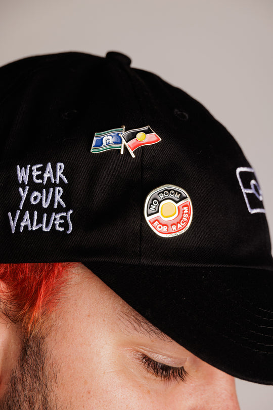 Clothing The Gaps. No Room For Racism Pin. Circle shaped pin with aboriginal flag inside and words 'No Room' in the black section of flag and 'For Racism' in red section of flag. Text is in gold colour with capital bold text.