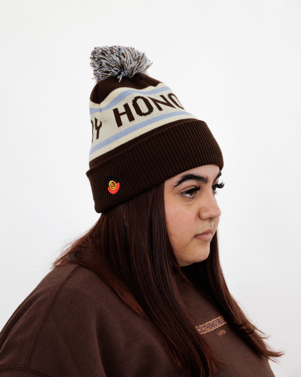 Clothing The Gaps. Honour Country Beanie. Chocolate brown base with thick butter cream middle section and two skinny ice blue lines above and below the chocolate brown text 'Honour Country.' Chocolate brown, butter cream and ice blue pom pom on top of beanie.