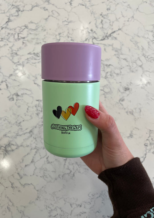 Clothing The Gaps. Reusable Stainless steel and ceramic base frank green mug. Light pastel purple lid. Pastel mint green base with image of 3 Hearts that are coloured  black, yellow and red. The colours of the aboriginal flag. Under the hearts Clothing The Gaps written in bubble writing underneath in black. 