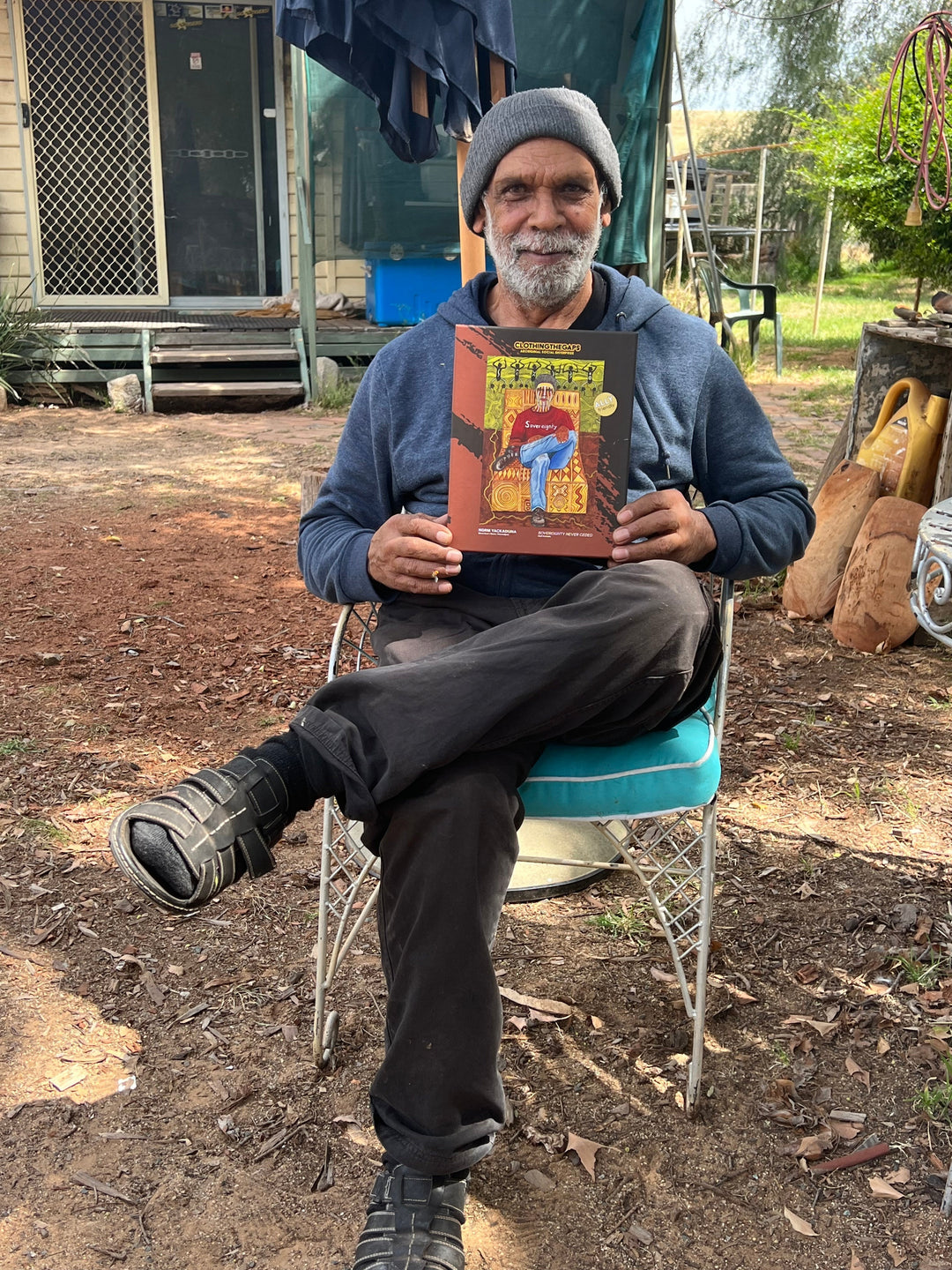 Clothing The Gaps. 1000 piece Puzzle of Aboriginal man with traditional face paint wearing a red sovereignty t-shirt on a yellow possum skin cloak chair. With a brown and green aboriginal style background using dots lines to tell story. Artwork by Norm Yakaduna.