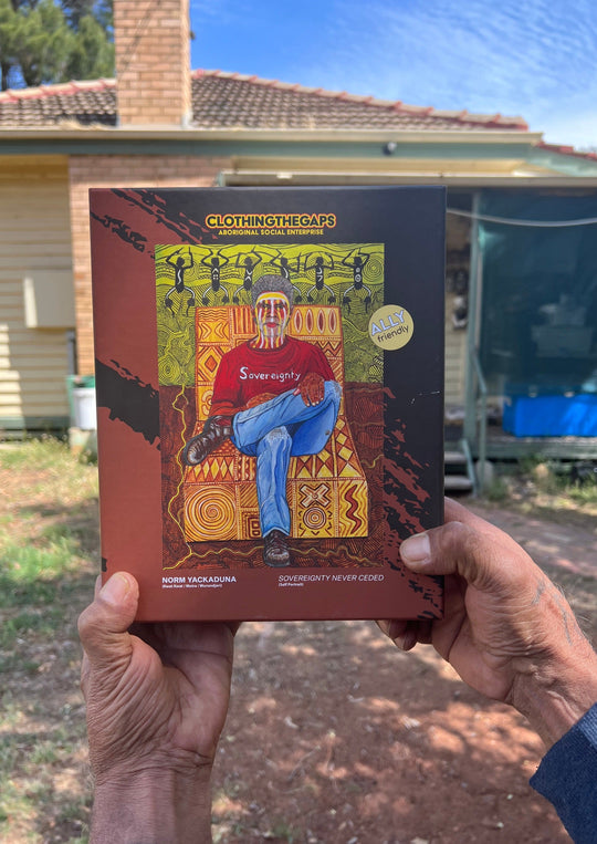 Clothing The Gaps. 1000 piece Puzzle of Aboriginal man with traditional face paint wearing a red sovereignty t-shirt on a yellow possum skin cloak chair. With a brown and green aboriginal style background using dots lines to tell story. Artwork by Norm Yakaduna.