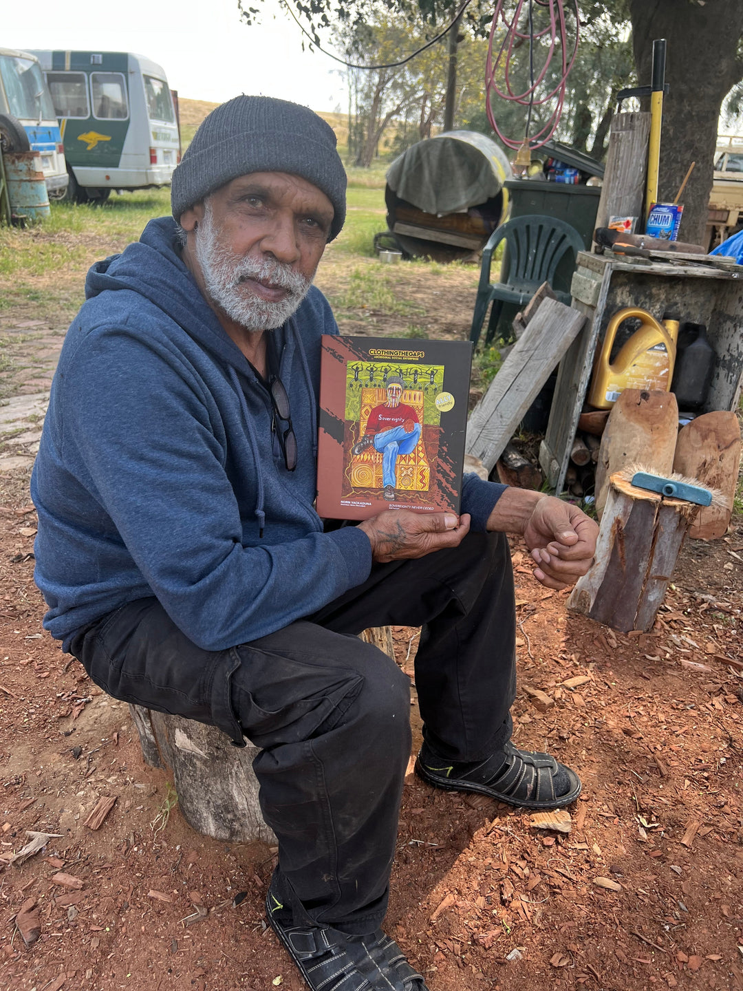 Clothing The Gaps. 1000 piece Puzzle of Aboriginal man with traditional face paint wearing a red sovereignty t-shirt on a yellow possum skin cloak chair. With a brown and green aboriginal style background using dots lines to tell story. Artwork by Norm Yakaduna.