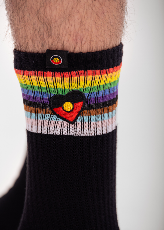 Black sock with rainbow striped cuff featuring a Aboriginal flag heart design on a white background