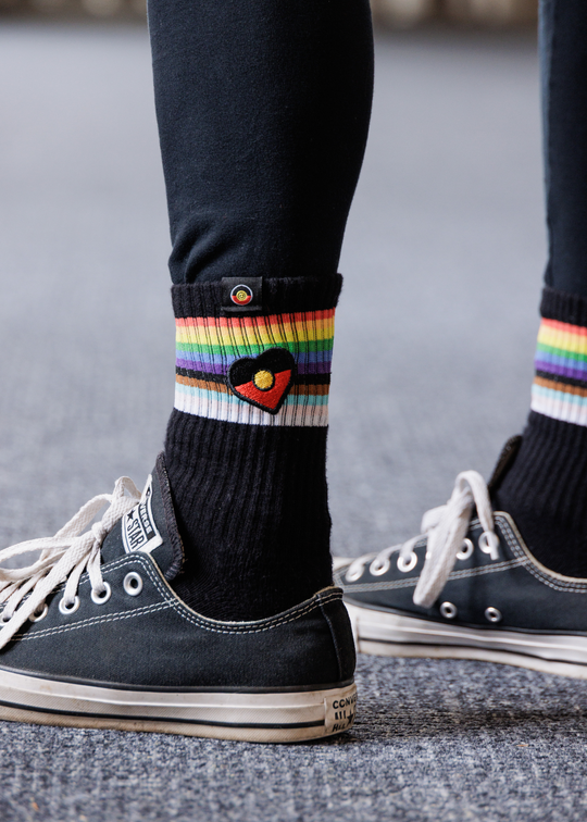 Person wearing black Clothing The Gaps socks with rainbow heart design and white sneakers on a gray pavement background
